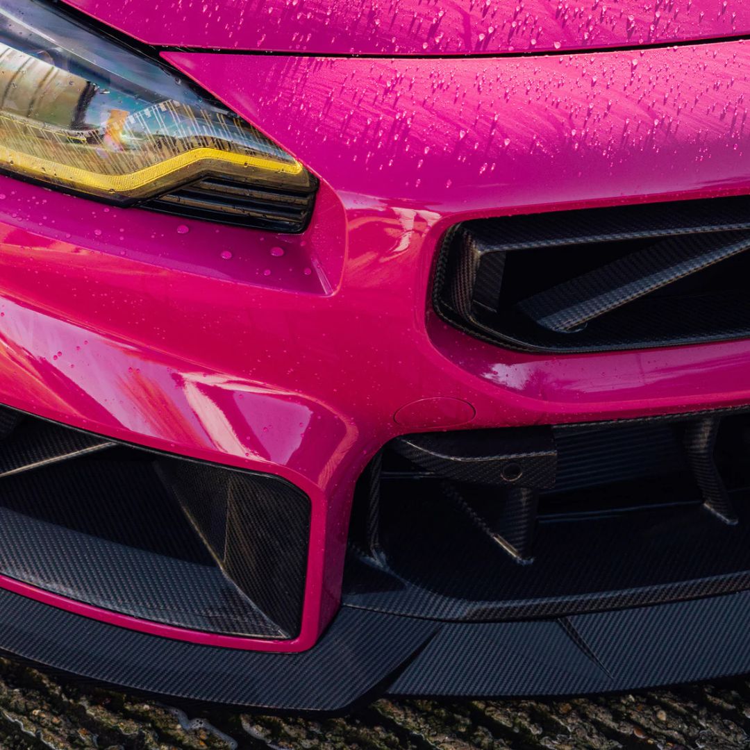 Close-up of a pink BMW M2 front bumper with water droplets on a textured surface.