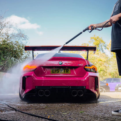 Person washing a pink BMW M2 with a pressure washer on a sunny day.