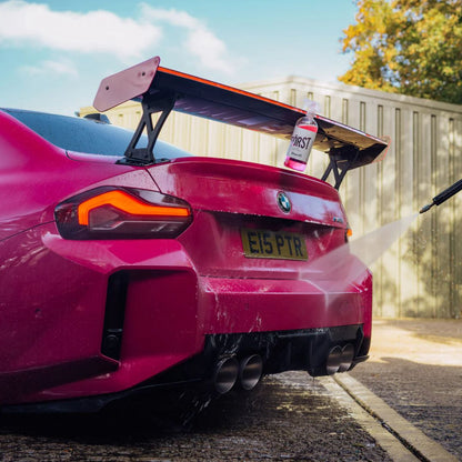 Pink BMW M2 car being washed with a soap bottle labeled 'Stjarnagloss Forst' on a sunny day.