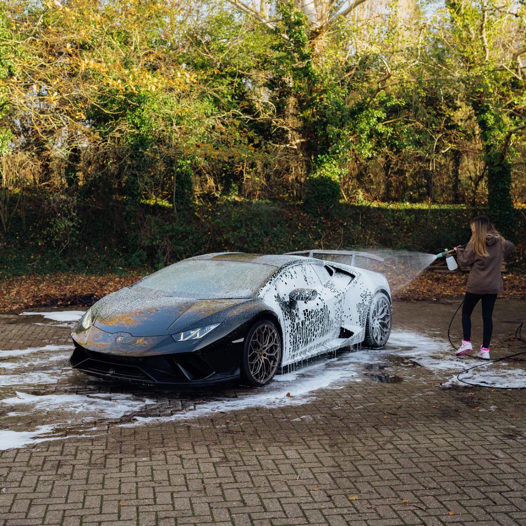 Person washing a black Lamborghini car with soap suds on a driveway.