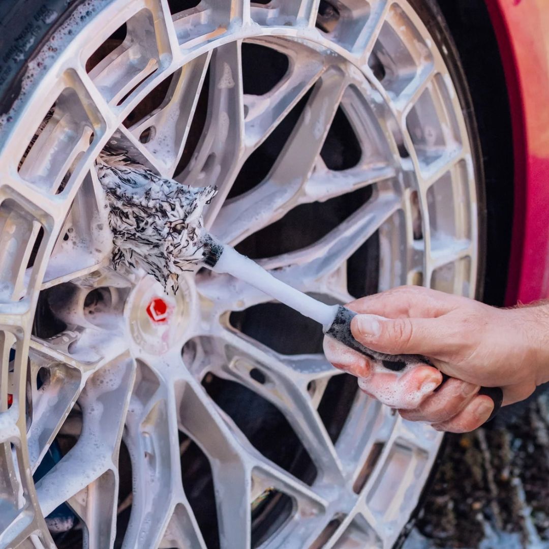 A close-up of a hand using a foam-covered Stjarnagloss Gravling - Microfibre Wheel Brush to clean a silver multi-spoke car wheel.