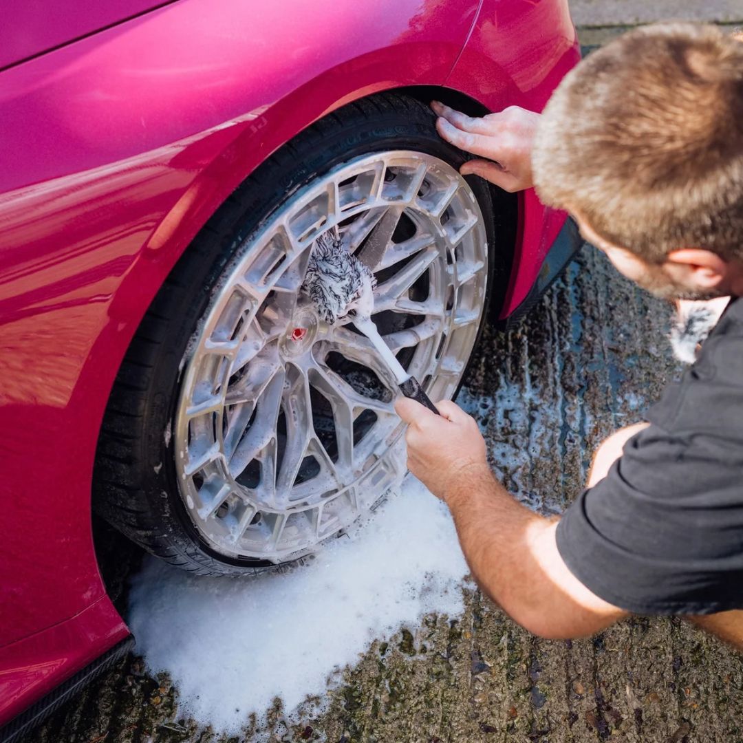 A man washes a silver alloy wheel of a bright pink car with soapy water on a wet pavement surface.