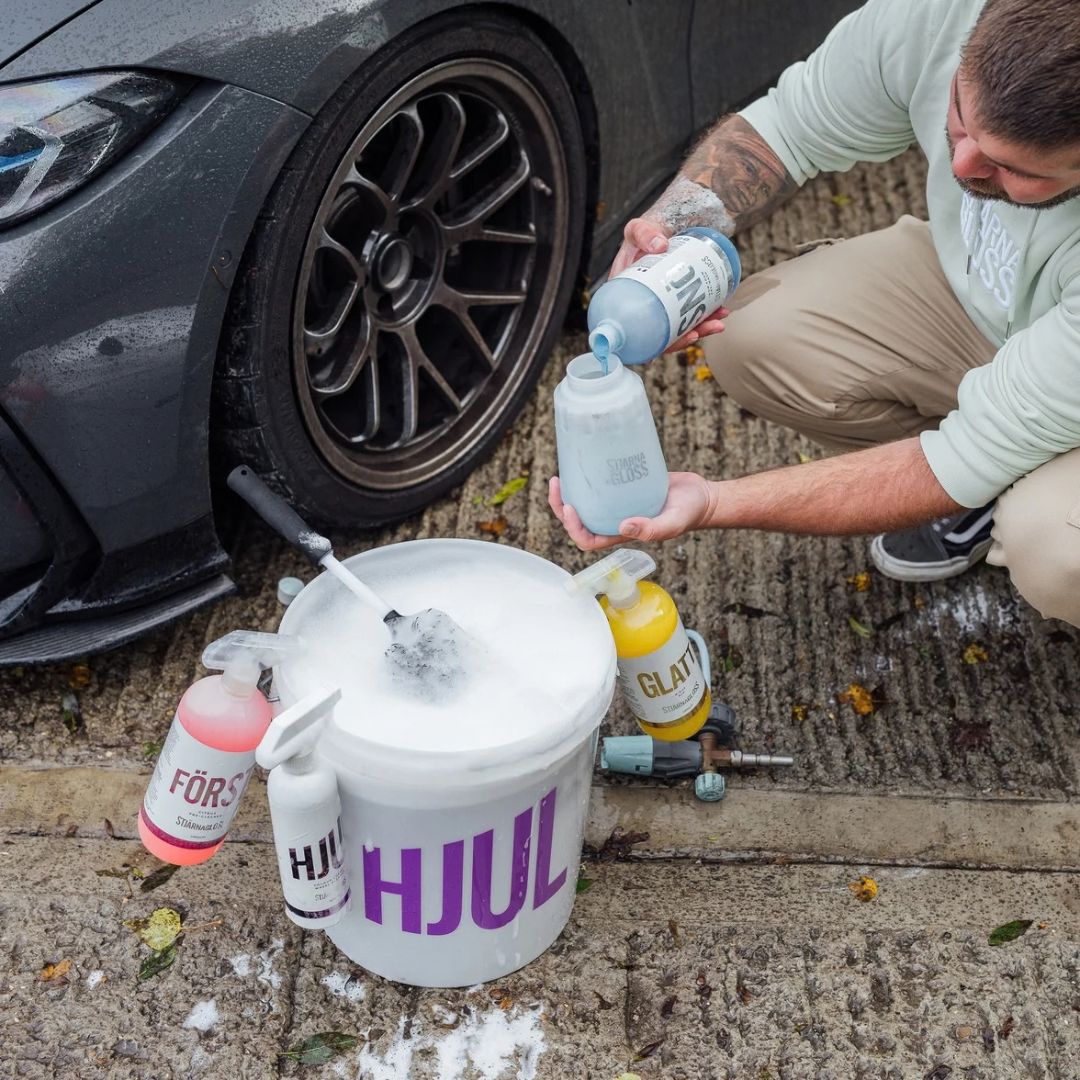 A man kneels beside a dark car wheel, pouring liquid into a white bucket filled with foam while cleaning supplies, including bottles and a drill, sit on the wet pavement. The bucket has large purple 'HJUL' lettering.