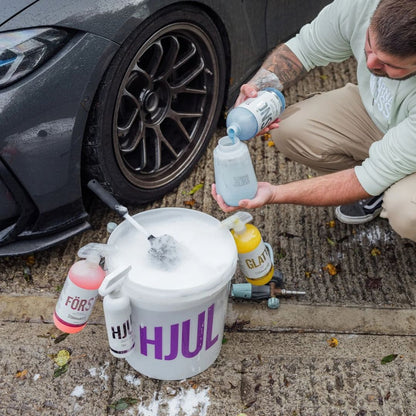 A man kneels beside a dark car wheel, pouring liquid into a white bucket filled with foam while cleaning supplies, including bottles and a drill, sit on the wet pavement. The bucket has large purple 'HJUL' lettering.