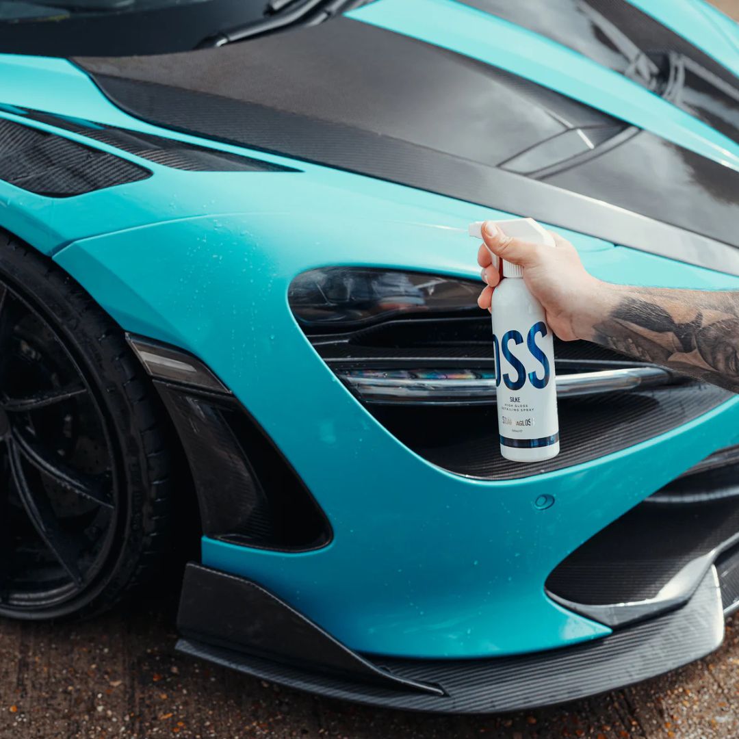 Person holding a bottle labeled 'Stjarnagloss Silke' against a turquoise sports car McLaren