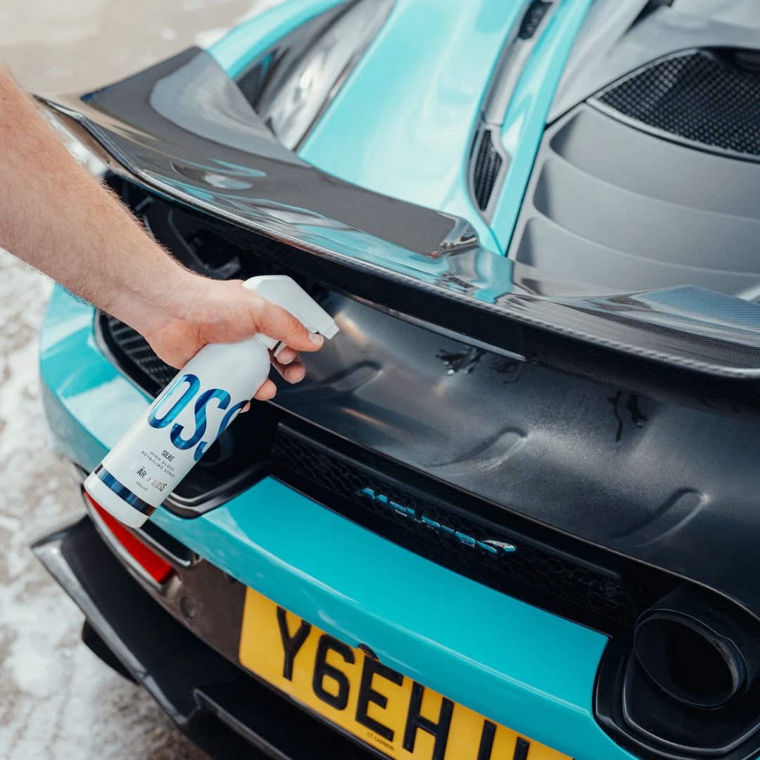 Person using a spray bottle labeled 'Stjarnagloss Silke' on a turquoise McLaren with a visible license plate.