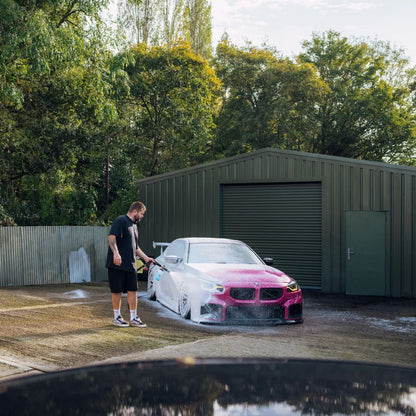 Person washing a pink BMW M2 with Stjarnagloss Sno - pH-Neutral Snow Foam  outdoors with trees and a shed in the background