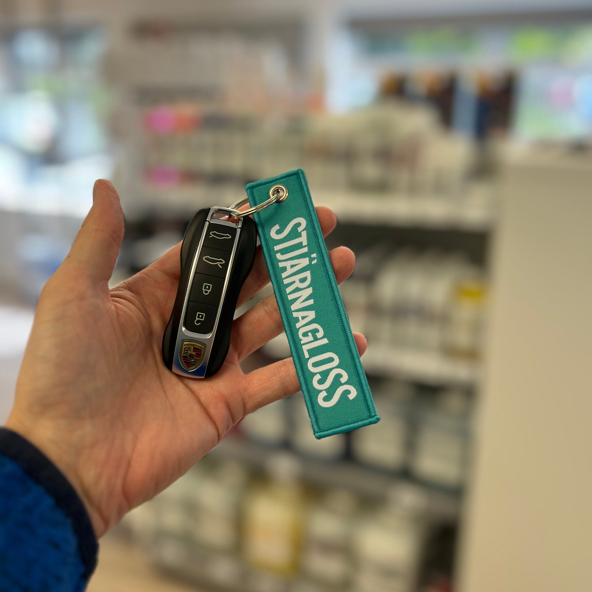 Hand holding a Stjarnagloss Porsche key fob with a 'Stjärnagloss' tag in a store setting