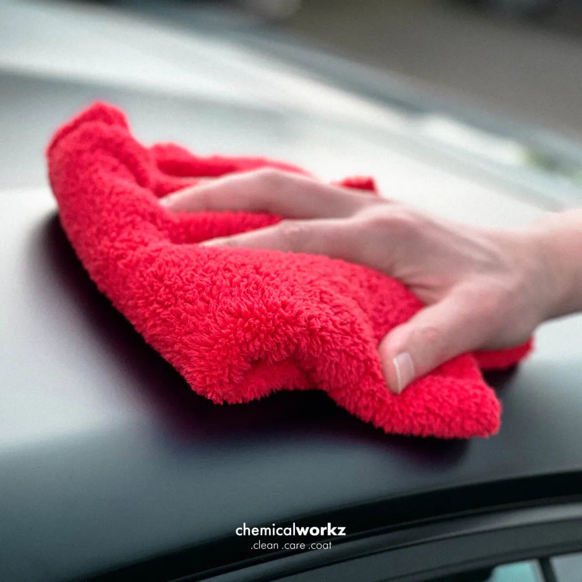 A close-up of a hand wiping a dark car surface with a bright red microfibere 600gsm cloth. The text 'chemicalworkz clean care coat' is visible at the bottom.