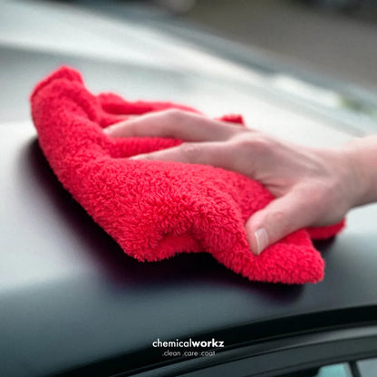 A close-up of a hand wiping a dark car surface with a bright red microfibere 600gsm cloth. The text 'chemicalworkz clean care coat' is visible at the bottom.