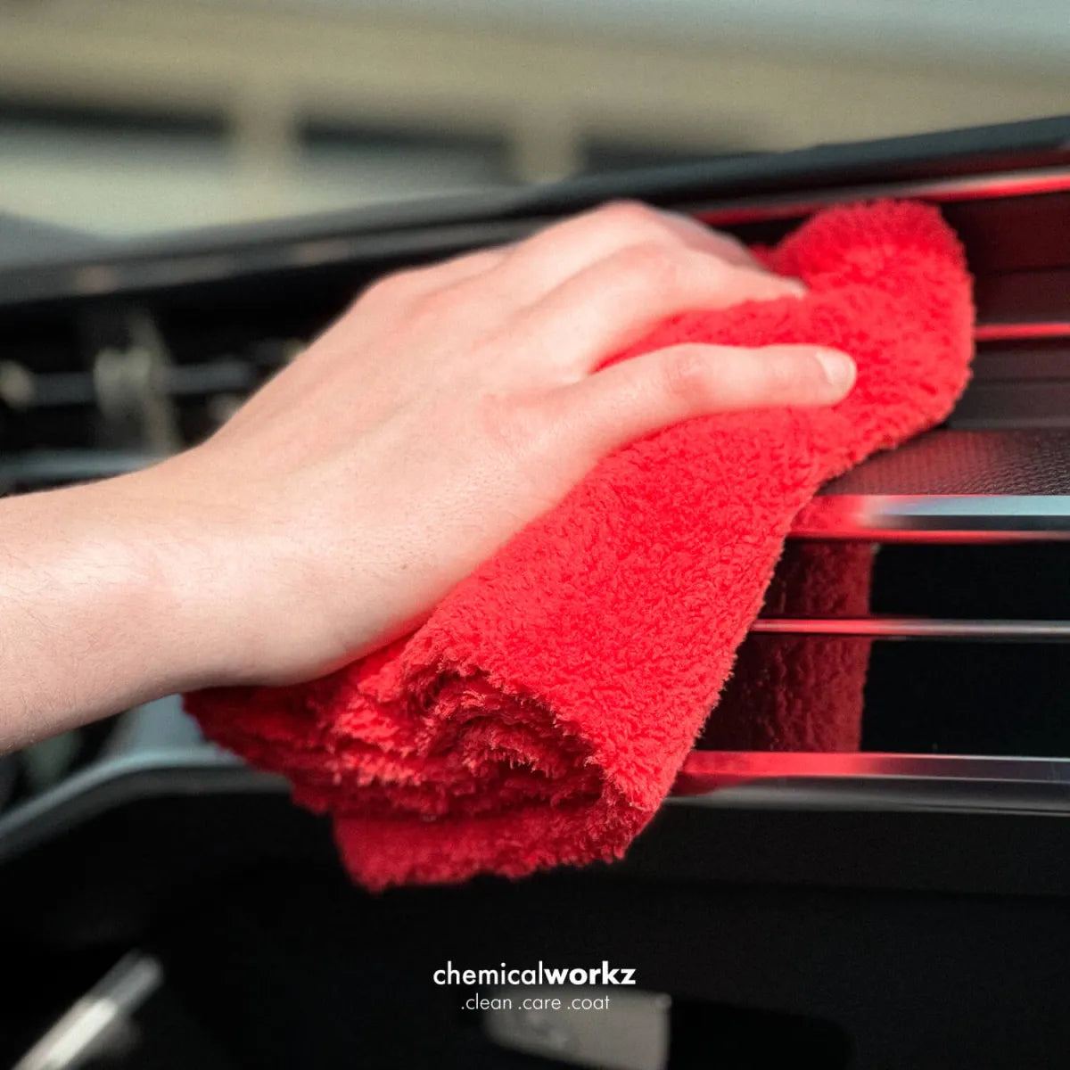 A close-up of a hand wiping a car dashboard vent with a bright red microfiber cloth. The brand name 'chemicalworkz' is visible at the bottom of the image.