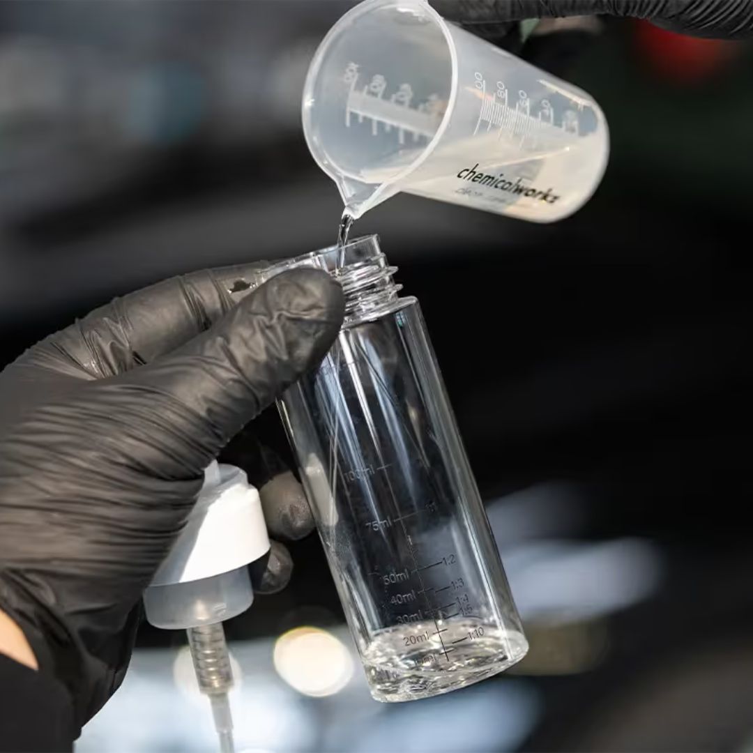 Person pouring liquid from a measuring cup into a clear container with measurement markings, wearing black gloves.